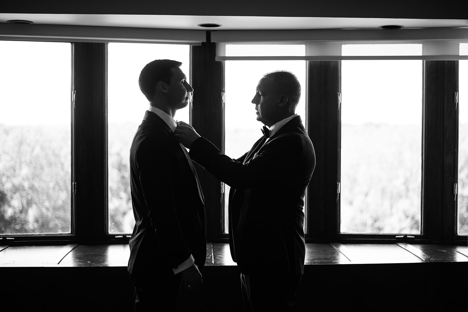 Black and white silhouette of the groom's father adjusting his bow tie in front of large windows.